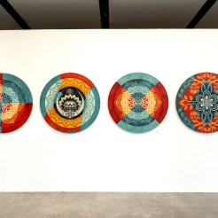 Four circular art pieces with intricate mandala-like patterns in red, blue, yellow, and black, displayed on a white gallery…