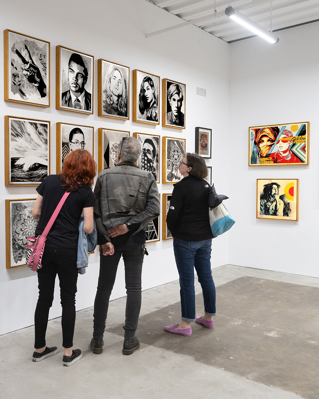 Three people view a gallery wall of framed black and white prints and colorful artwork.