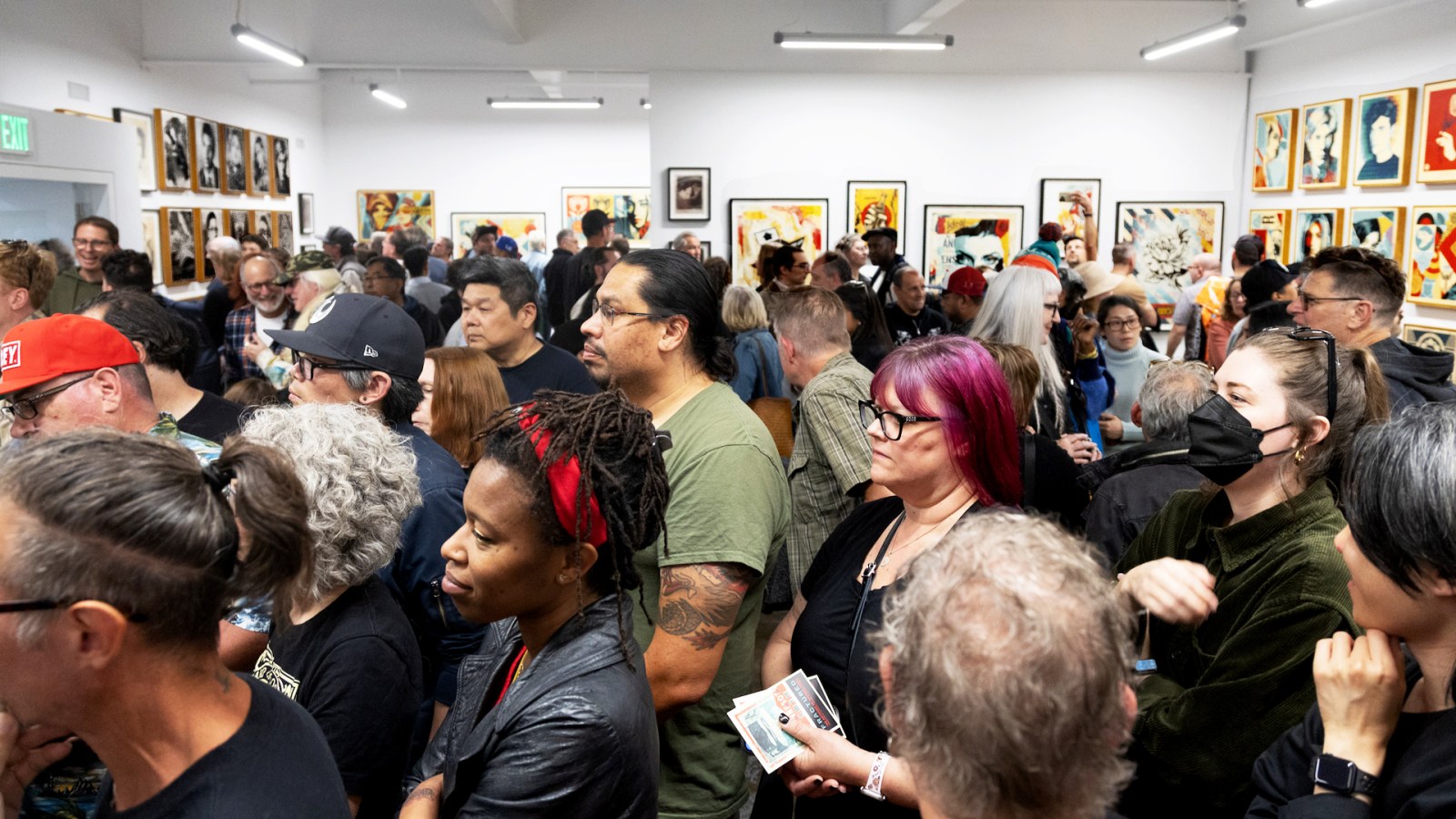 Crowded art gallery opening; diverse attendees view artwork on walls.
