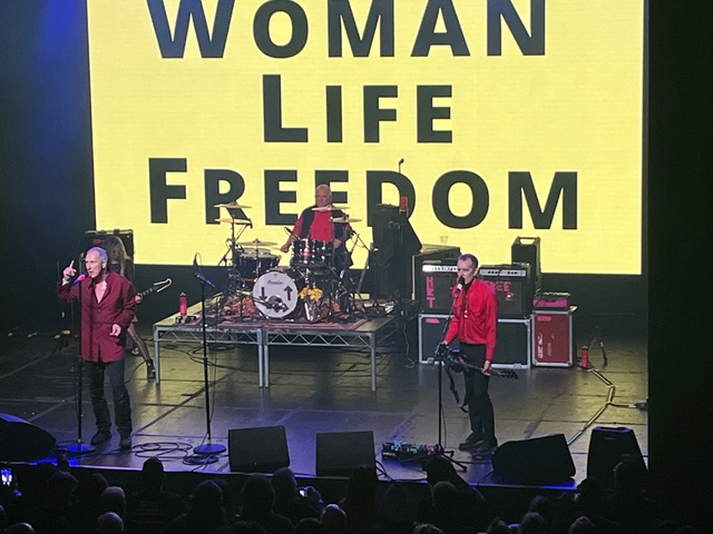 A band performs on stage with a large yellow screen behind them displaying the text: "WOMAN LIFE FREEDOM".