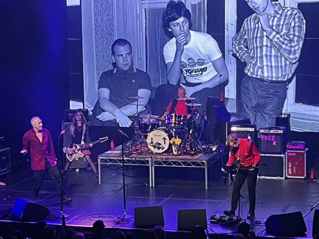 A band performs on stage with large blue-tinted images of young men in the background.