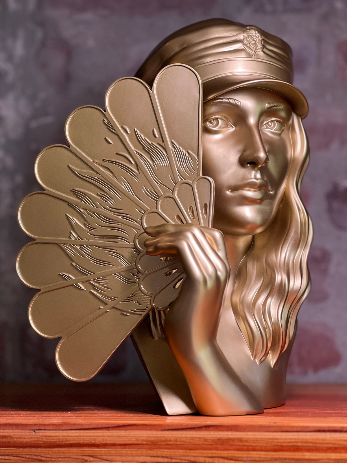 Gold bust of a woman wearing a cap, holding an ornate fan.