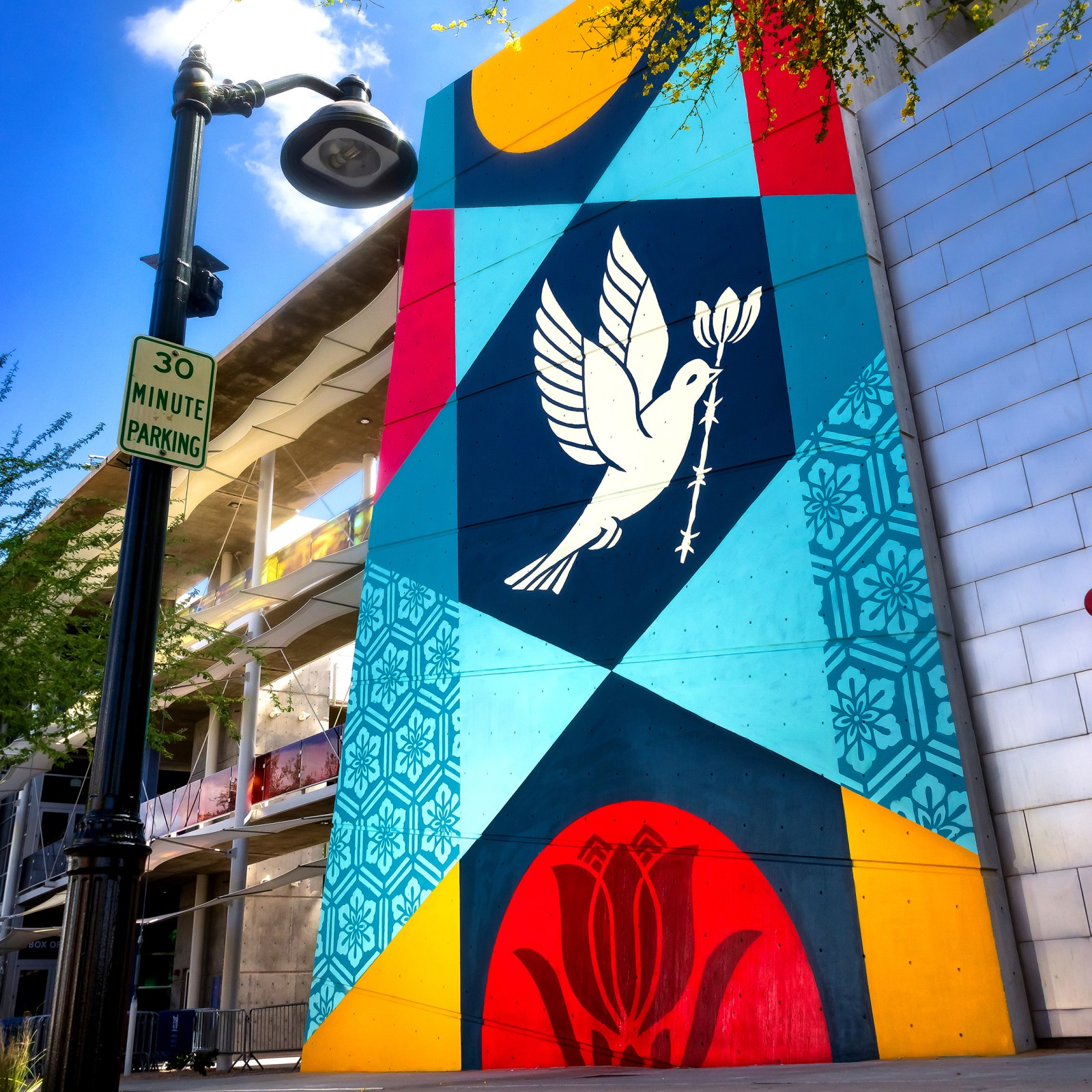A vibrant mural on a building featuring geometric patterns and a white dove holding a branch, in red, yellow, blue, and…
