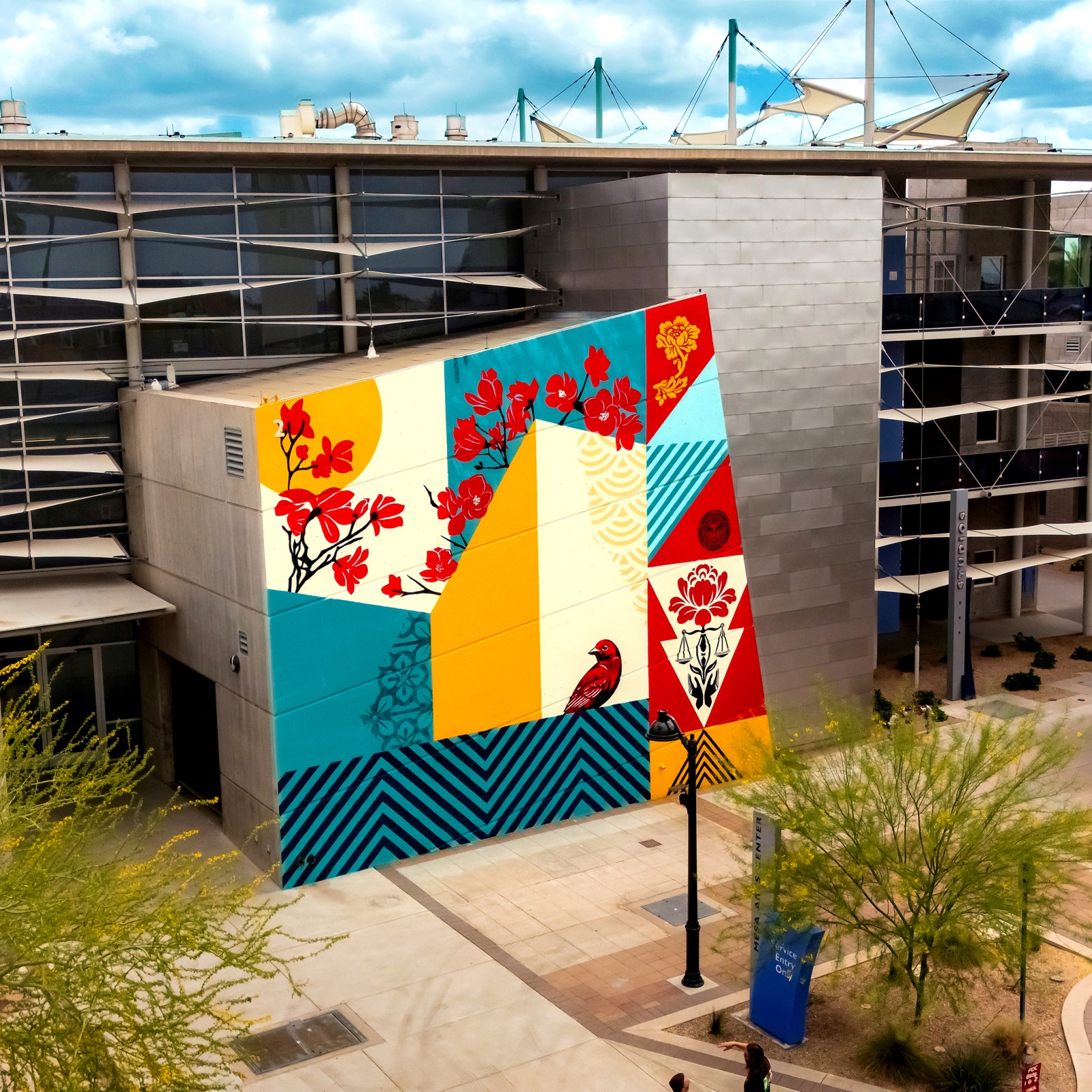 An aerial view of a vibrant mural on a building featuring geometric patterns, red flowers, and a bird, in red, yellow, blue…