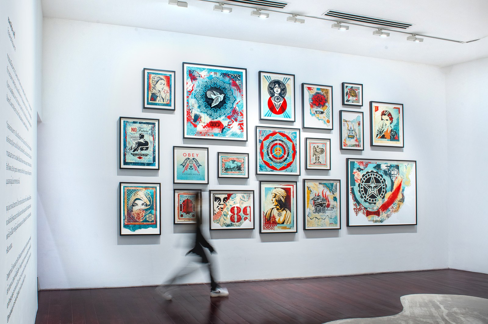 A gallery wall filled with various framed artworks, mostly colorful prints, with a blurred person walking by.