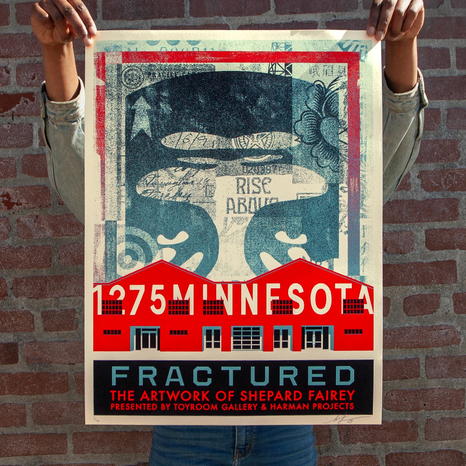 A person holds a poster for 'Fractured: The Artwork of Shepard Fairey.' It features two red buildings, '1275 Minnesota,' and&hellip;