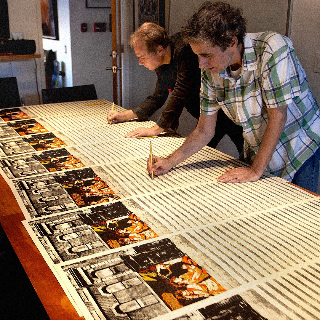 Two men, Shepard Fairey and Glen E. Friedman, signing a long table covered with