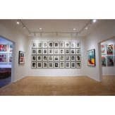 Art gallery with light wood floors, white walls, and a large grid of small framed black and white portraits.