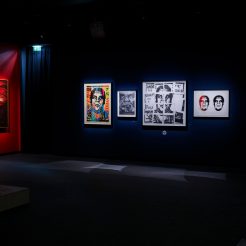 A dimly lit art gallery with several framed artworks on the walls, including portraits and abstract designs, and a red…