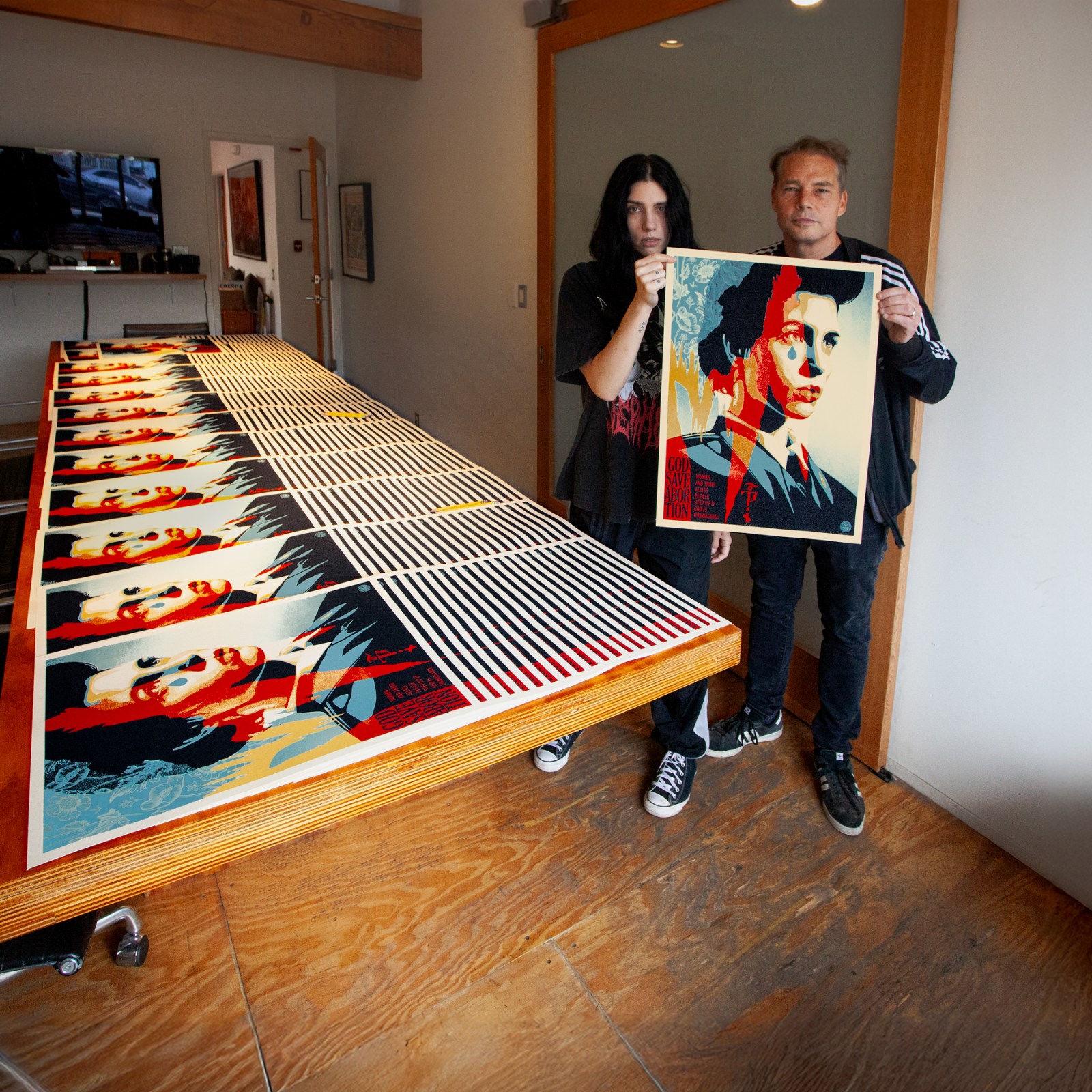 A man and woman hold up a poster of a woman with a tear, while a long table of identical prints stretches behind them.