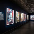 An art exhibition hallway with framed artworks on dark blue walls, including a large portrait of a woman with a flower in…
