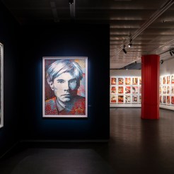 An art exhibition room with framed artworks on dark blue walls, including portraits of women and Andy Warhol, and a red…
