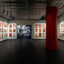 An art exhibition room with walls covered in framed artworks, mostly red and white, and a central red column.
