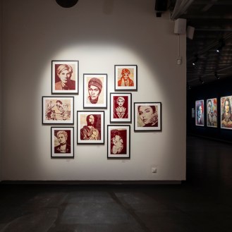 A gallery wall with nine framed art prints, mostly portraits in red and black tones. To the right, a dark hallway leads to…