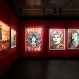 A gallery wall painted red, featuring four framed art prints. The first reads "MAKE ART NOT WAR" with a woman's face. The…