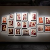 A gallery wall with a large grid of framed art prints, mostly portraits and landscapes in red and black tones, illuminated…