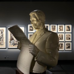 A white bust of a man with a skull face, wearing a suit and reading a newspaper, stands in a gallery. Framed art prints line…