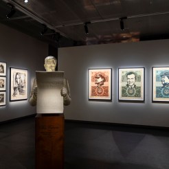 A white bust of a man with a skull face, wearing a suit and reading a newspaper, stands in a gallery. Framed art prints line…