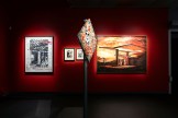 A red gallery wall with a framed black and white artwork of a building interior on the left. A tall, diamond-shaped…
