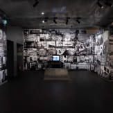 A gallery room with walls covered in black and white collages of faces and urban scenes. A television and a ramp are in the…