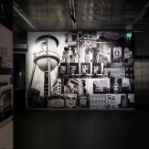 A gallery room with a large black and white mural featuring a water tower and collages of faces and urban scenes. An…