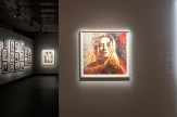 A gallery space with a large framed portrait of a woman with closed eyes and a flower in her hair, illuminated on a white…