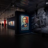 A gallery space with a large framed portrait of Barack Obama with the word "HOPE" on a dark blue wall. People are visible in…