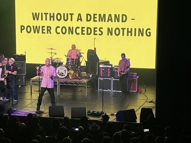 A band performs on stage with a large yellow screen behind them displaying the text: "WITHOUT A DEMAND - POWER CONCEDES&hellip;